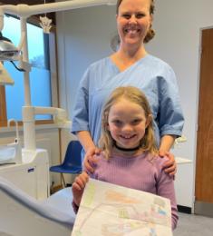 En kvindelig tandplejer holder om skuldrene på en pige med langt hår, der står lige foran hende. Pigen holder en tegning. Begge står ved siden af en tandlægestol.